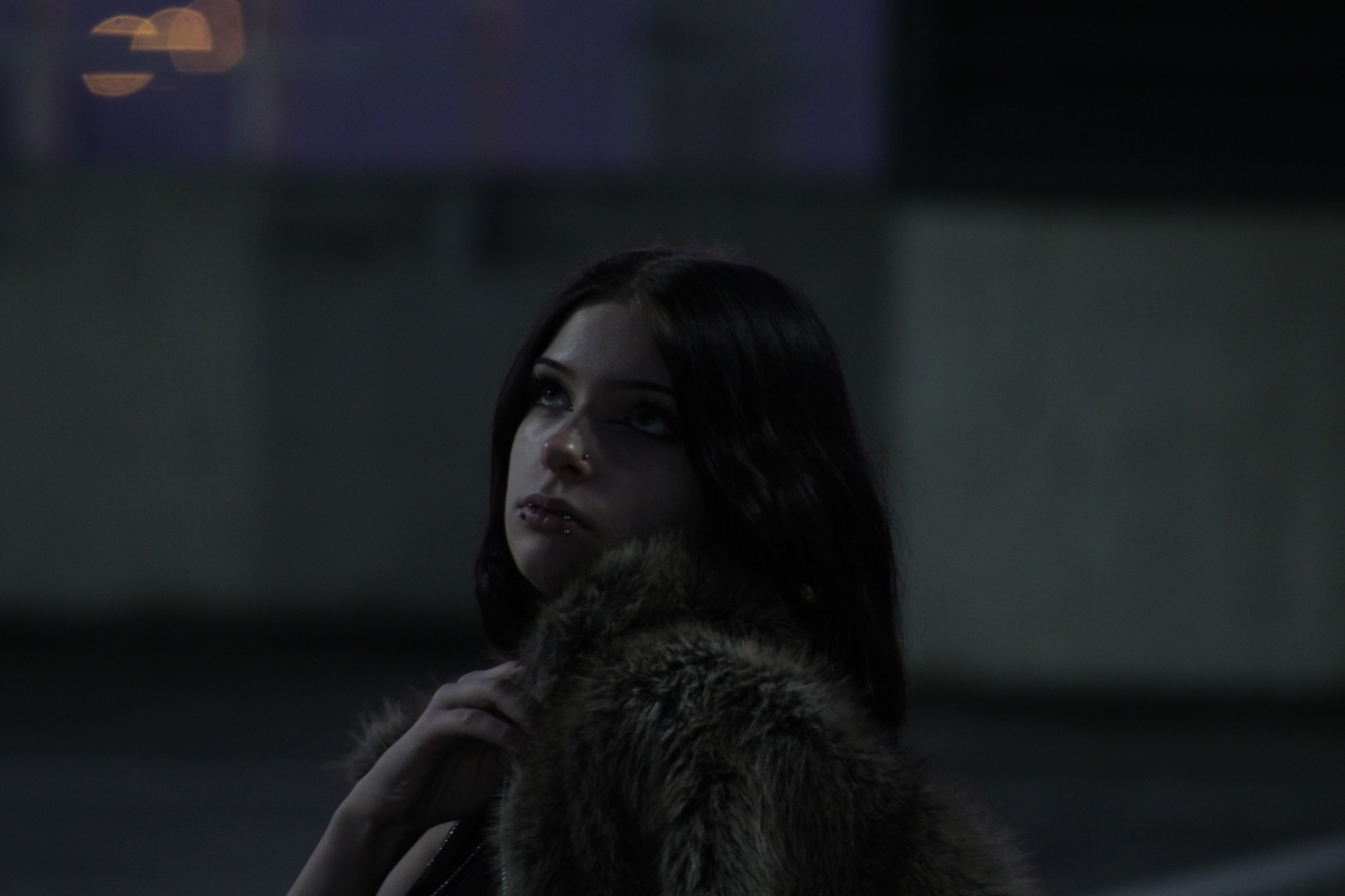 Portret kobiety z piercingiem, spoglądającej w górę, ubrana w futrzane okrycie na ciemnym tle.