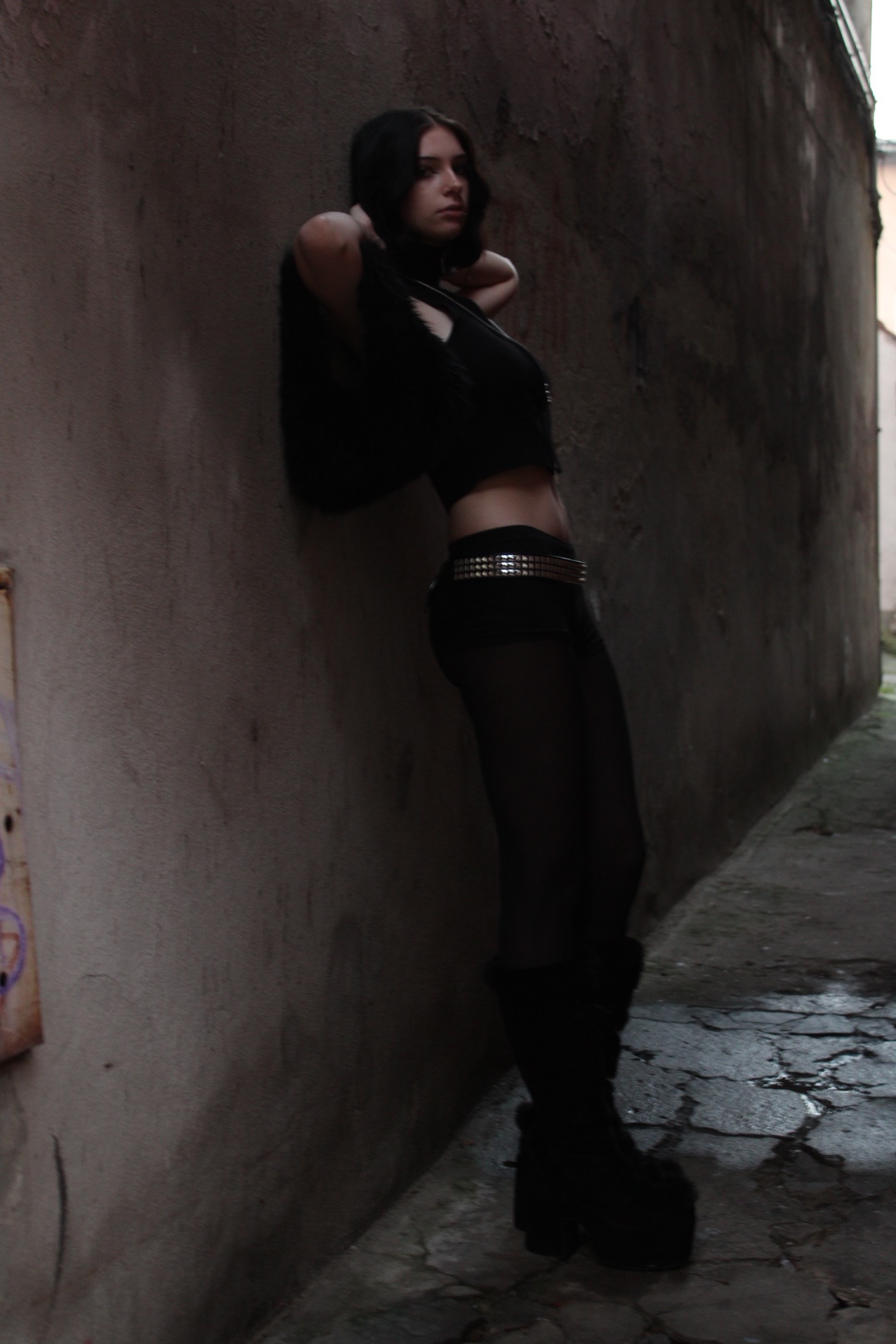 Młoda kobieta w czarnym stroju pozuje, opierając się o szarą ścianę w wąskiej uliczce, z widocznym paskiem z ćwiekami i masywnymi butami na platformie.