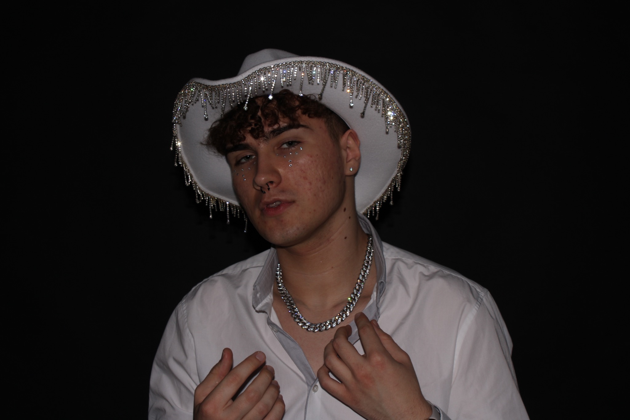 Młody mężczyzna w białym kapeluszu kowbojskim z frędzlami z kryształków, srebrnym łańcuchem na szyi i białej koszuli na czarnym tle.