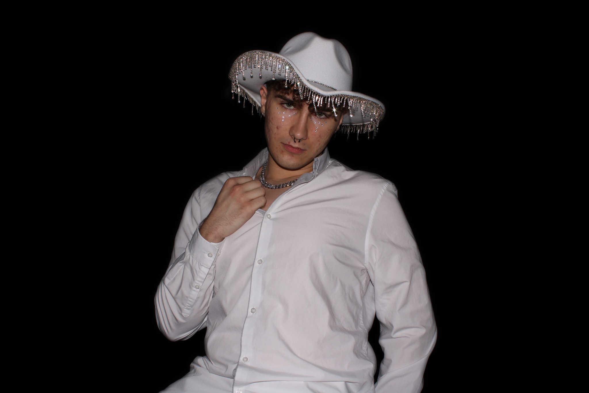Młody mężczyzna w białej koszuli i kapeluszu kowbojskim z frędzlami z kryształków, pozujący na czarnym tle.