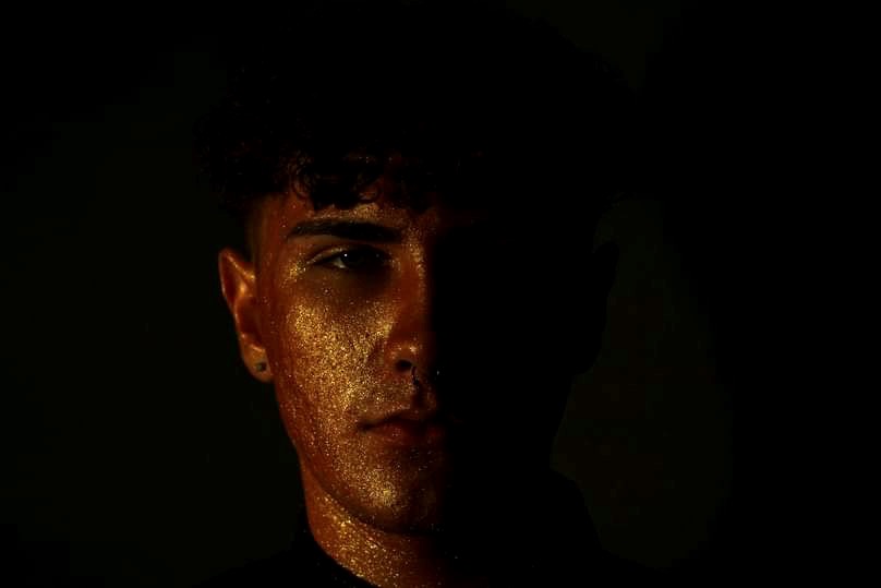 Portret młodego mężczyzny z połową twarzy w cieniu, oświetlonej złotym światłem, z kroplami potu lub brokatu na skórze.