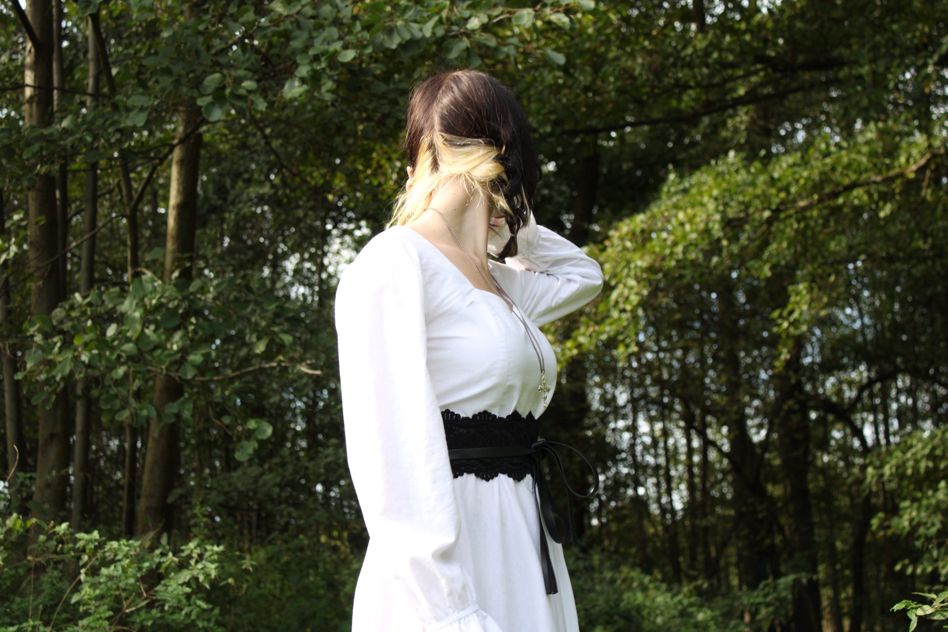 Kobieta w białej sukience z czarnym pasem pozuje na tle drzew w lesie, włosy z blond pasemkami, widok z boku.