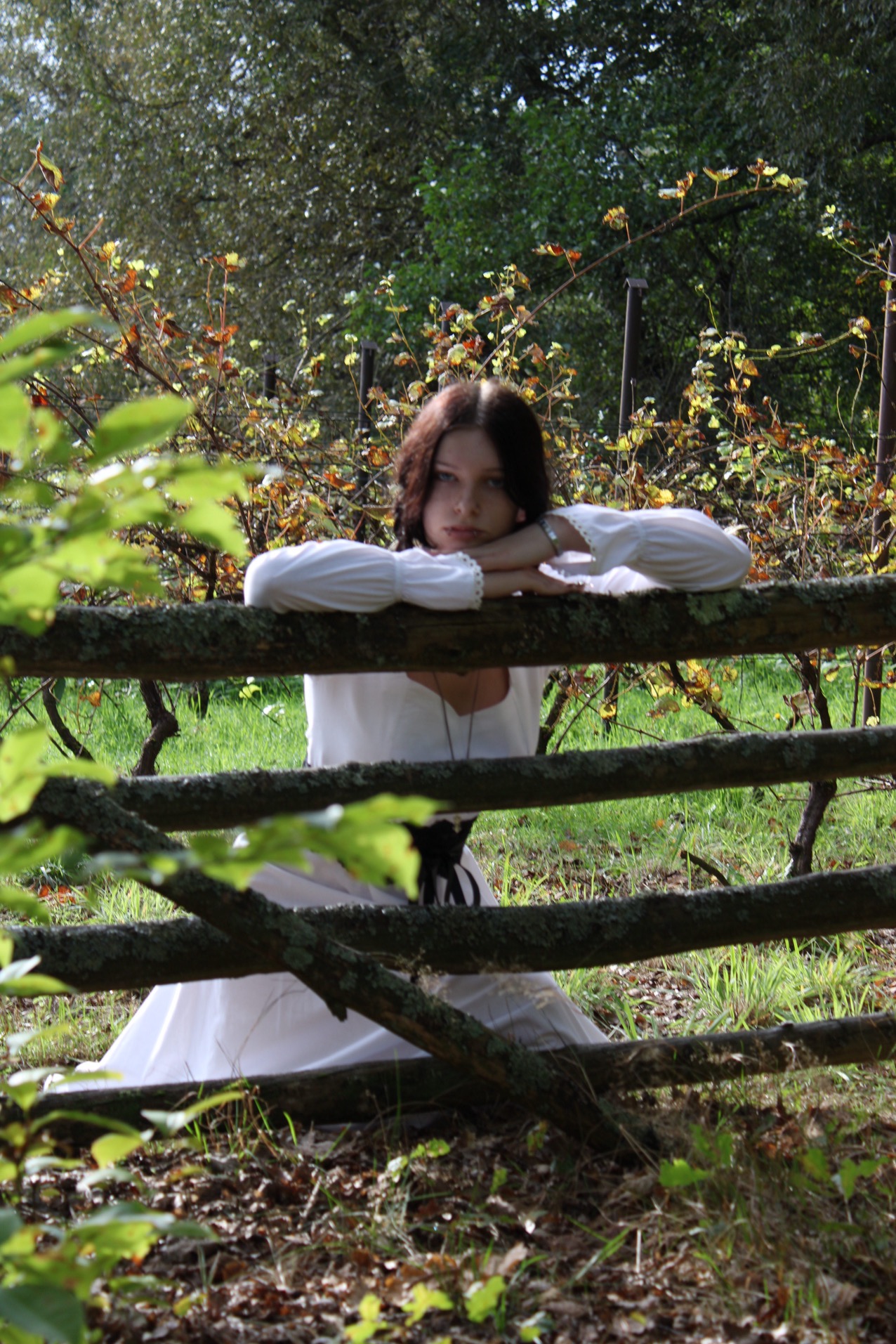 Młoda kobieta w białej sukni, opierająca się o stary, omszały drewniany płot na tle winnicy.