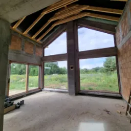 Surowy stan wnętrza domu z dużymi oknami z widokiem na łąkę i las, widoczne elementy konstrukcyjne, cegły, betonowe słupy i drewniane krokwie dachu, drabina oparta o ścianę, instalacja...