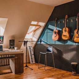 DANGER STUDIO DAWID ANGERHOEFER - Profesjonalne studio muzyczne z akustycznymi panelami na ścianie, czterema gitarami wiszącymi pod girlandą z żarówek, klawiaturą, monitorem z oprogramowaniem DAW, mikrofonem i bębnem djembe.