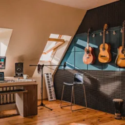 Profesjonalne studio muzyczne z akustycznymi panelami na ścianie, czterema gitarami wiszącymi pod girlandą z żarówek, klawiaturą, monitorem z oprogramowaniem DAW, mikrofonem i bębnem djembe.