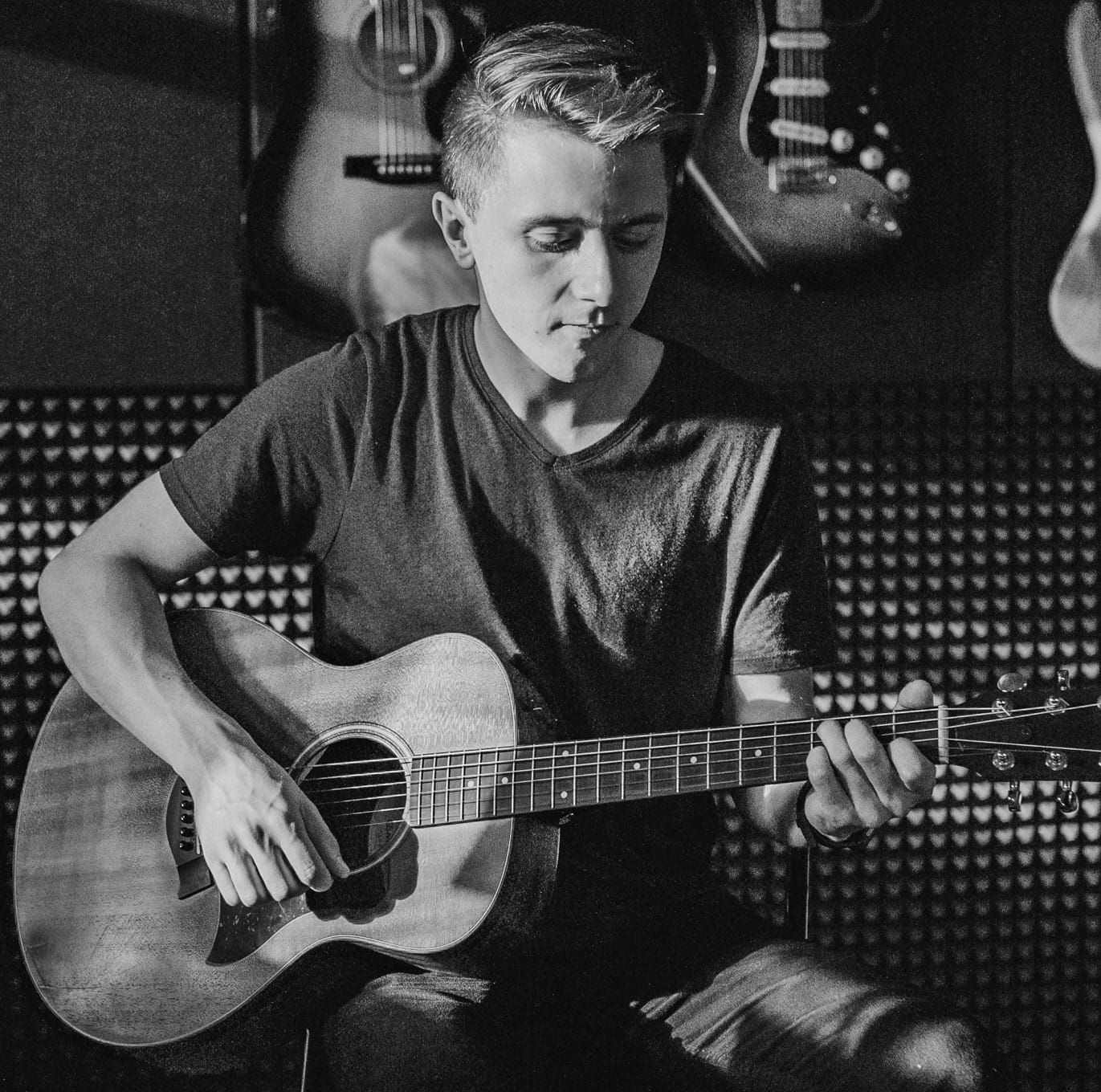 Młody mężczyzna gra na gitarze akustycznej w pomieszczeniu z panelami akustycznymi, w tle widoczne inne gitary.