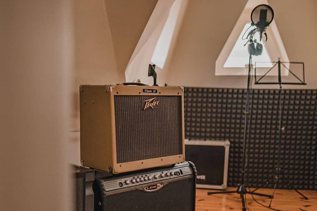 Wnętrze pomieszczenia z akustyczną pianką na ścianach, wzmacniacze gitarowe marki Peavey Classic 30 i Line 6, mikrofon pojemnościowy na statywie oraz pulpit na nuty.
