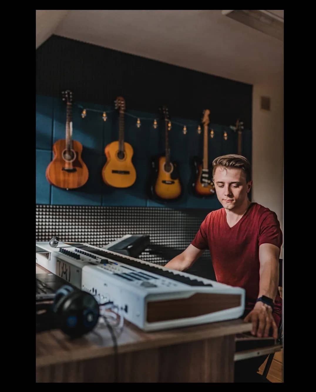 Młody mężczyzna gra na białym keyboardzie w profesjonalnie wyposażonym pomieszczeniu z akustycznymi panelami i wiszącymi gitarami podświetlonymi lampkami.
