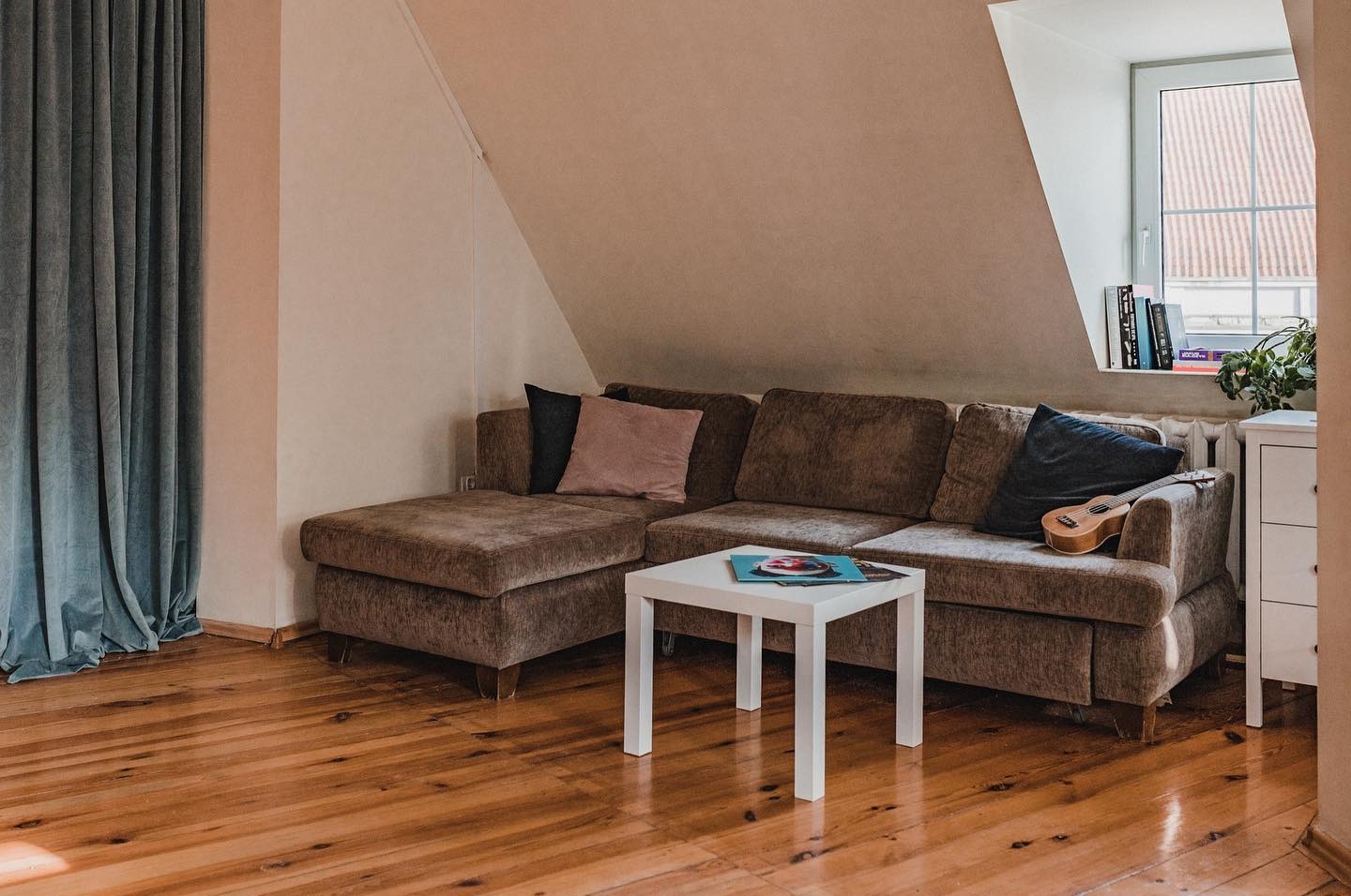 Ukulele spoczywa na brązowej sofie w jasnym pokoju w Olsztynie, obok okna z książkami i rośliną doniczkową.