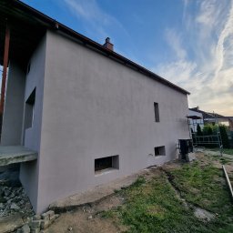 SUPERFLUM SPÓŁKA Z OGRANICZONĄ ODPOWIEDZIALNOŚCIĄ - Otynkowany dom jednorodzinny z szarą elewacją w trakcie wykańczania, widok z boku na tle błękitnego nieba z chmurami. Widoczne rusztowania i materiały budowlane.