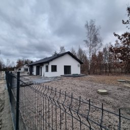 SUPERFLUM SPÓŁKA Z OGRANICZONĄ ODPOWIEDZIALNOŚCIĄ - Nowoczesny, parterowy dom z czarnym dachem i białą elewacją, widziany zza metalowego ogrodzenia na tle pochmurnego nieba i drzew.