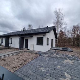 SUPERFLUM SPÓŁKA Z OGRANICZONĄ ODPOWIEDZIALNOŚCIĄ - Nowoczesny, parterowy dom z ciemną dachówką i kostką brukową. Widoczny etap prac wykończeniowych na zewnątrz budynku, z elementami przygotowania podłoża.
