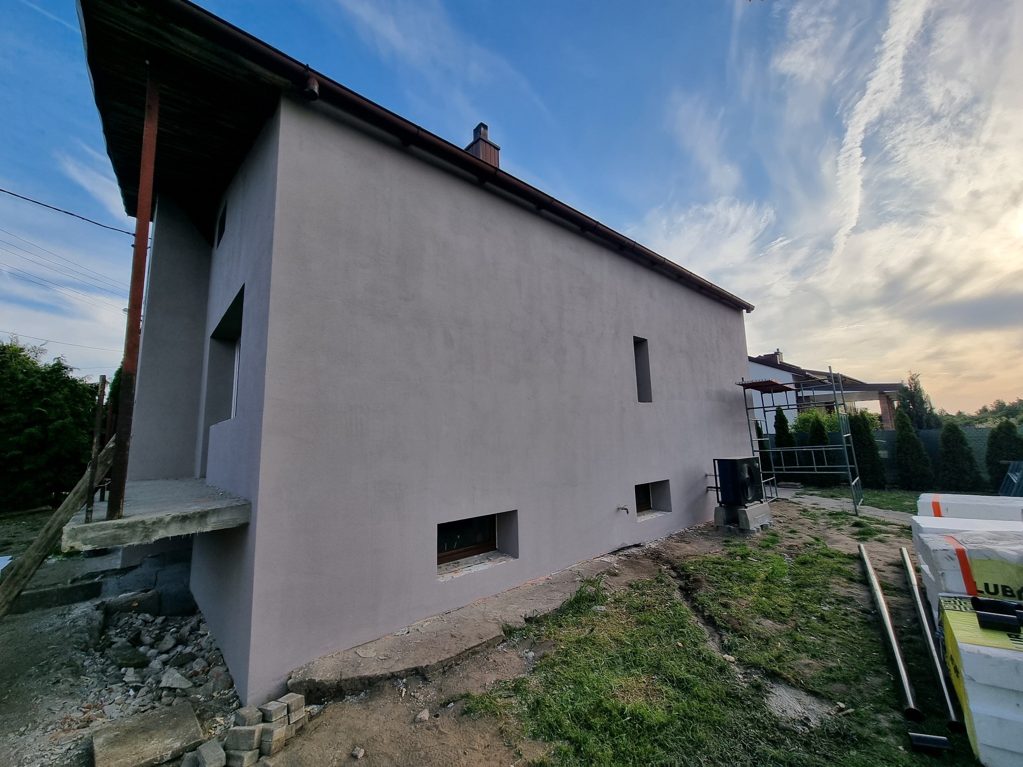 Otynkowany dom jednorodzinny z szarą elewacją w trakcie wykańczania, widok z boku na tle błękitnego nieba z chmurami. Widoczne rusztowania i materiały budowlane.