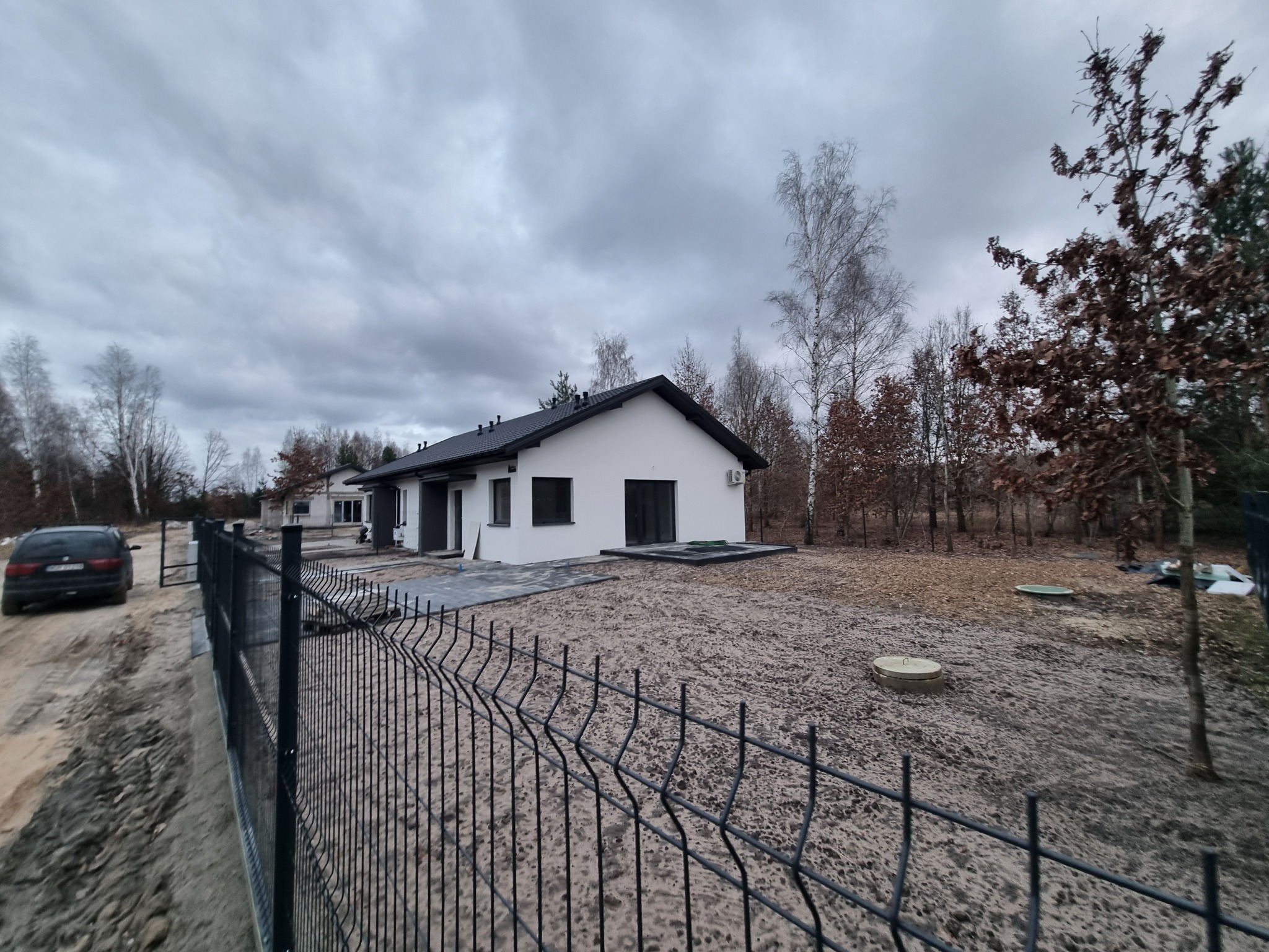 Nowoczesny, parterowy dom z czarnym dachem i białą elewacją, widziany zza metalowego ogrodzenia na tle pochmurnego nieba i drzew.