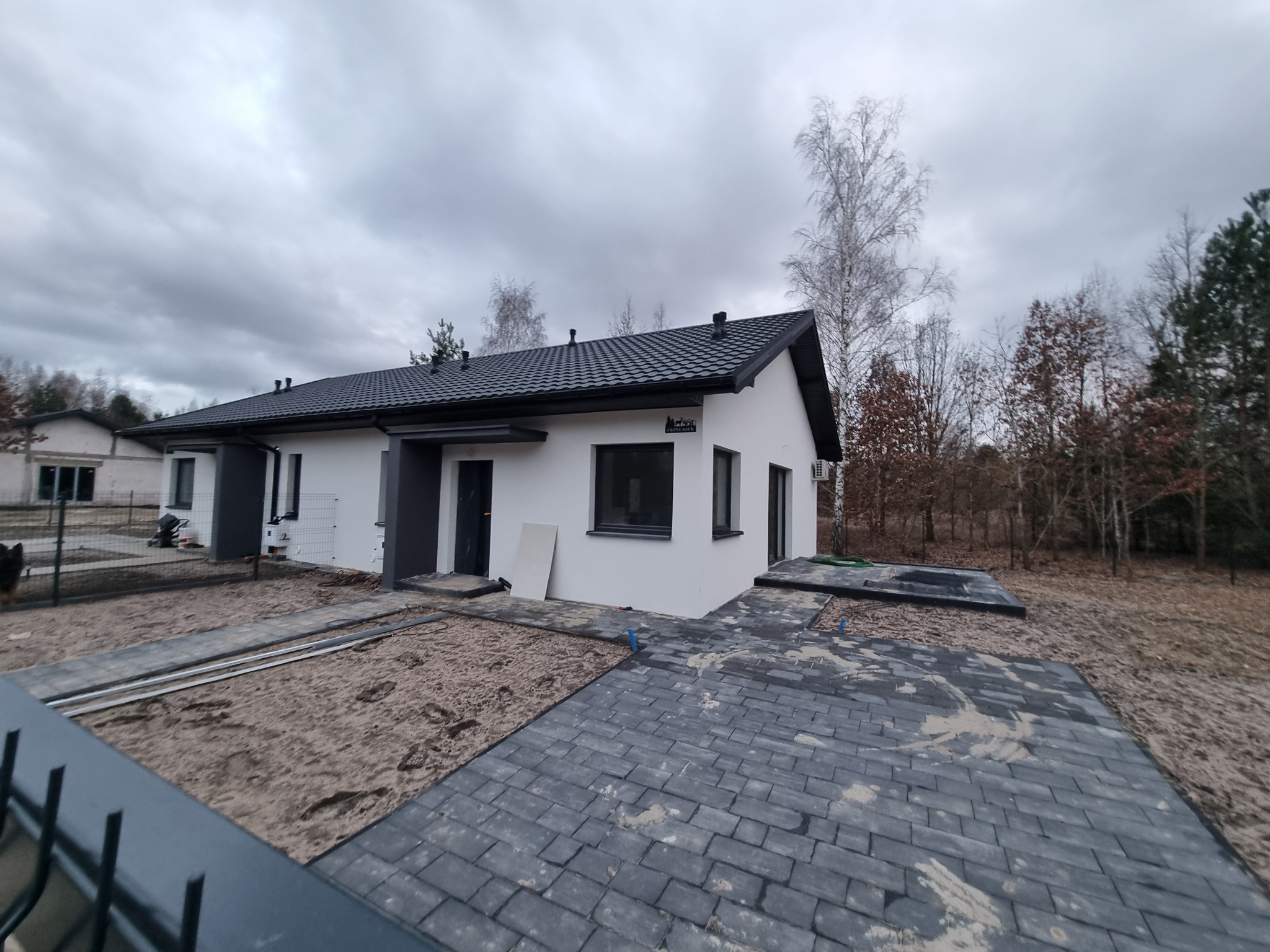 Nowoczesny, parterowy dom z ciemną dachówką i kostką brukową. Widoczny etap prac wykończeniowych na zewnątrz budynku, z elementami przygotowania podłoża.
