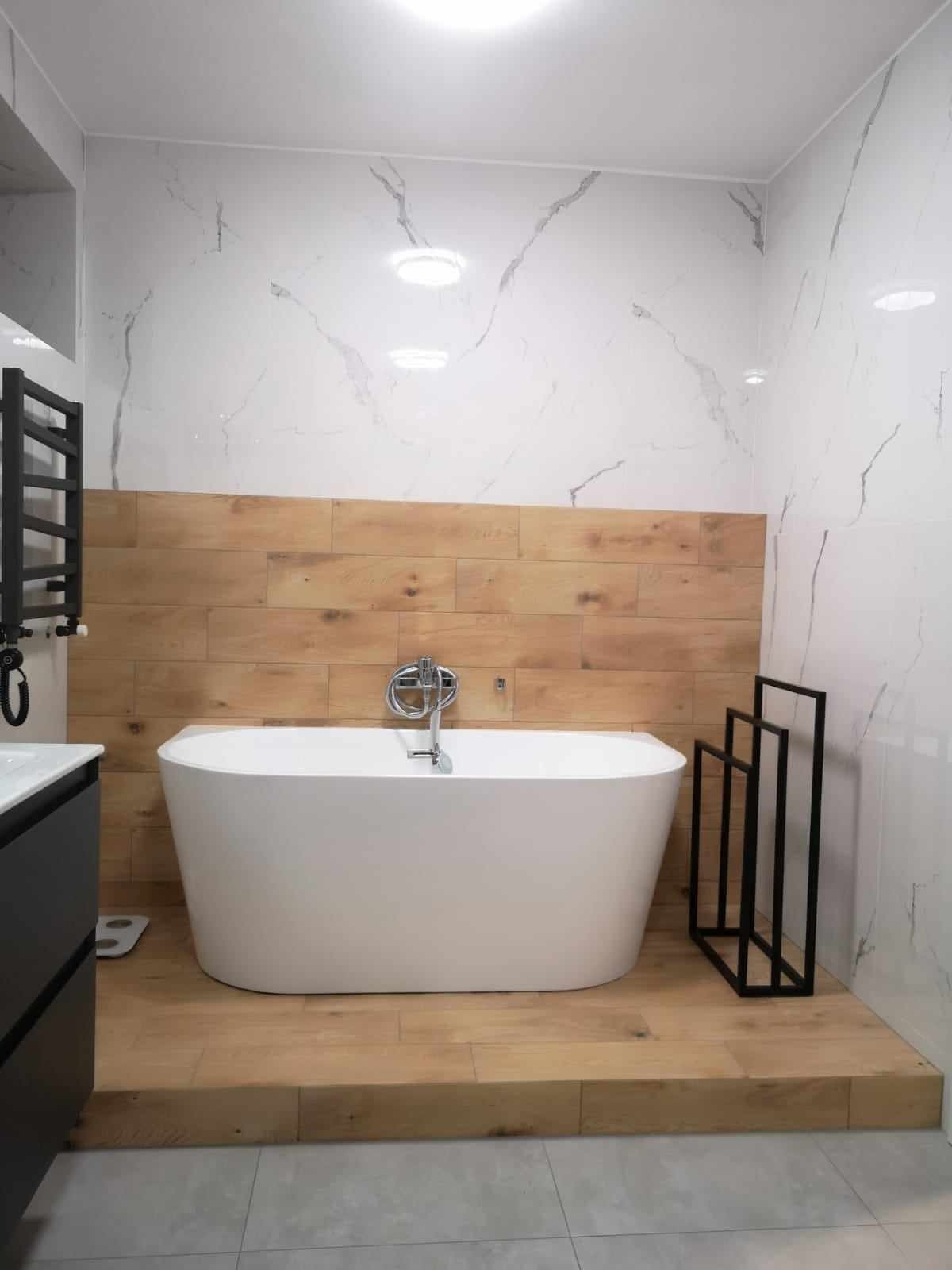 Nowoczesna łazienka z wolnostojącą wanną na drewnianym podeście, marmurowe płytki na ścianach i dekoracyjne drewniane panele. Minimalistyczny design z czarnymi akcentami.