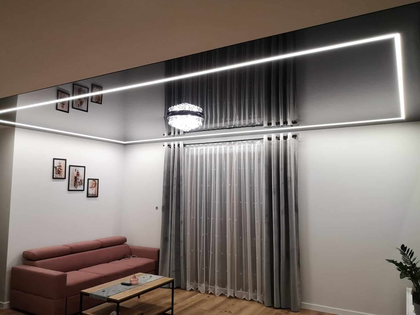 Nowoczesny salon z podwieszanym sufitem, oświetleniem LED w kształcie prostokąta i dekoracyjnymi zasłonami. Na ścianie galeria ramek ze zdjęciami rodzinnymi.