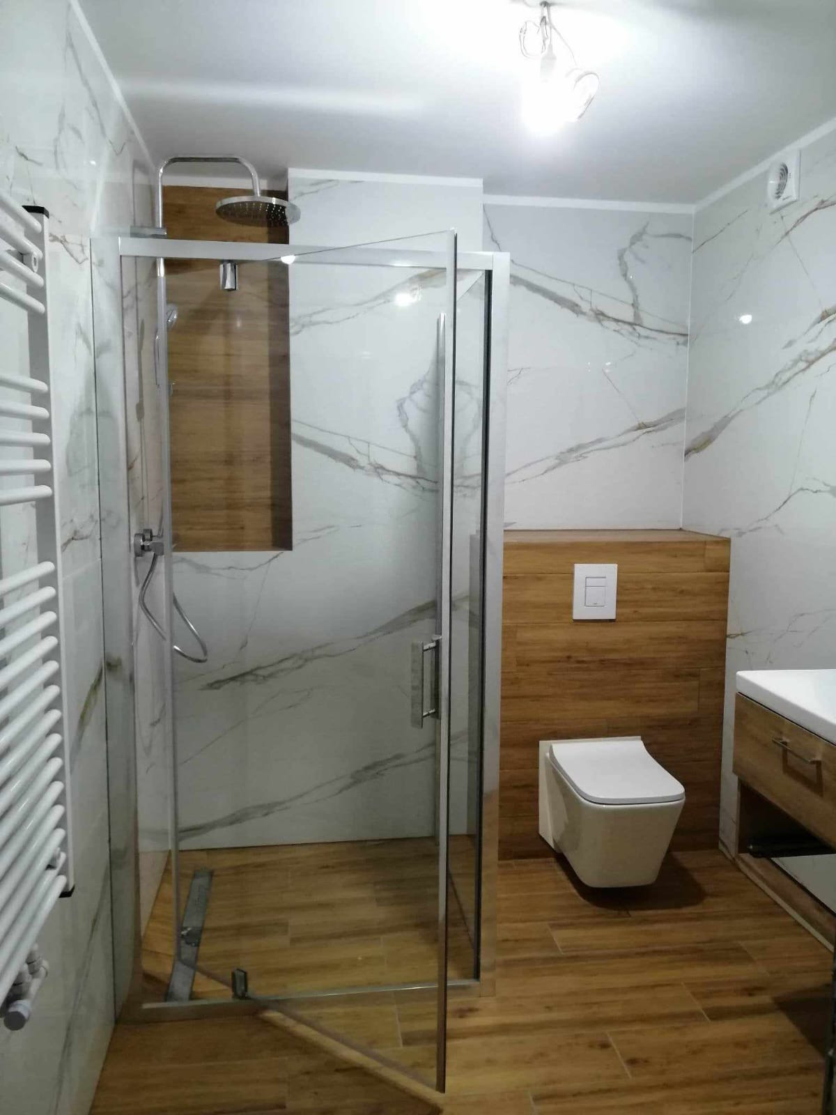 Nowoczesna łazienka po remoncie: kabina prysznicowa ze szklanymi drzwiami, marmurowe płytki z delikatnym wzorem i drewnopodobna zabudowa toalety.
