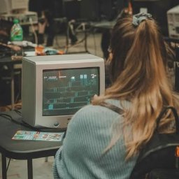 WARSZAWSKIE MUZEUM KOMPUTERÓW I GIER SPÓŁKA Z OGRANICZONĄ ODPOWIEDZIALNOŚCIĄ - Kobieta gra w grę retro na starym monitorze podczas wydarzenia z grami. W tle widoczni inni gracze i monitory. Atmosfera nostalgii i rywalizacji.