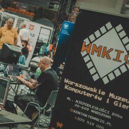 WARSZAWSKIE MUZEUM KOMPUTERÓW I GIER SPÓŁKA Z OGRANICZONĄ ODPOWIEDZIALNOŚCIĄ - Stoisko Warszawskiego Muzeum Komputerów i Gier na evencie, z osobami grającymi w gry na komputerach. Widoczny baner z logo muzeum w stylu pixel art.