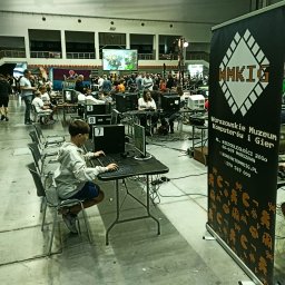 WARSZAWSKIE MUZEUM KOMPUTERÓW I GIER SPÓŁKA Z OGRANICZONĄ ODPOWIEDZIALNOŚCIĄ - Chłopiec gra na komputerze na evencie Warszawskiego Muzeum Komputerów i Gier, banner z logo, tłum ludzi w tle, numery stanowisk.