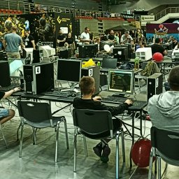 WARSZAWSKIE MUZEUM KOMPUTERÓW I GIER SPÓŁKA Z OGRANICZONĄ ODPOWIEDZIALNOŚCIĄ - Grupa osób uczestniczy w turnieju gier komputerowych, siedząc przy stanowiskach z monitorami. W tle widoczna publiczność i banery reklamowe sponsorów wydarzenia.