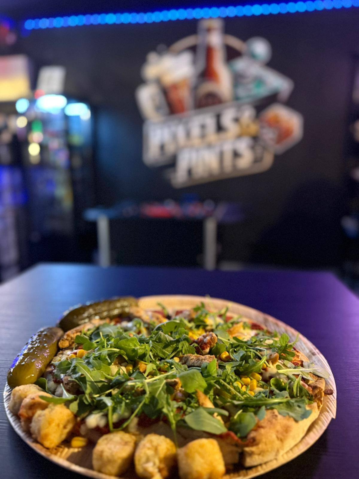 Pizza z rukolą, kukurydzą i kawałkami mięsa na drewnianym talerzu, z boku dwa ogórki konserwowe i smażone kulki, w tle logo 'Pixels & Pints' i automat z napojami.