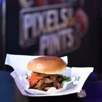 Apetyczny burger z wołowiną, pomidorami i rukolą, podany na białej papierowej tacce. W tle widoczny neon z nazwą 'PIXELS & PINTS'.