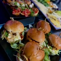 Mini burgery na czarnym talerzu z sałatą i wykałaczkami, w tle bruschetta z pomidorami i ser.