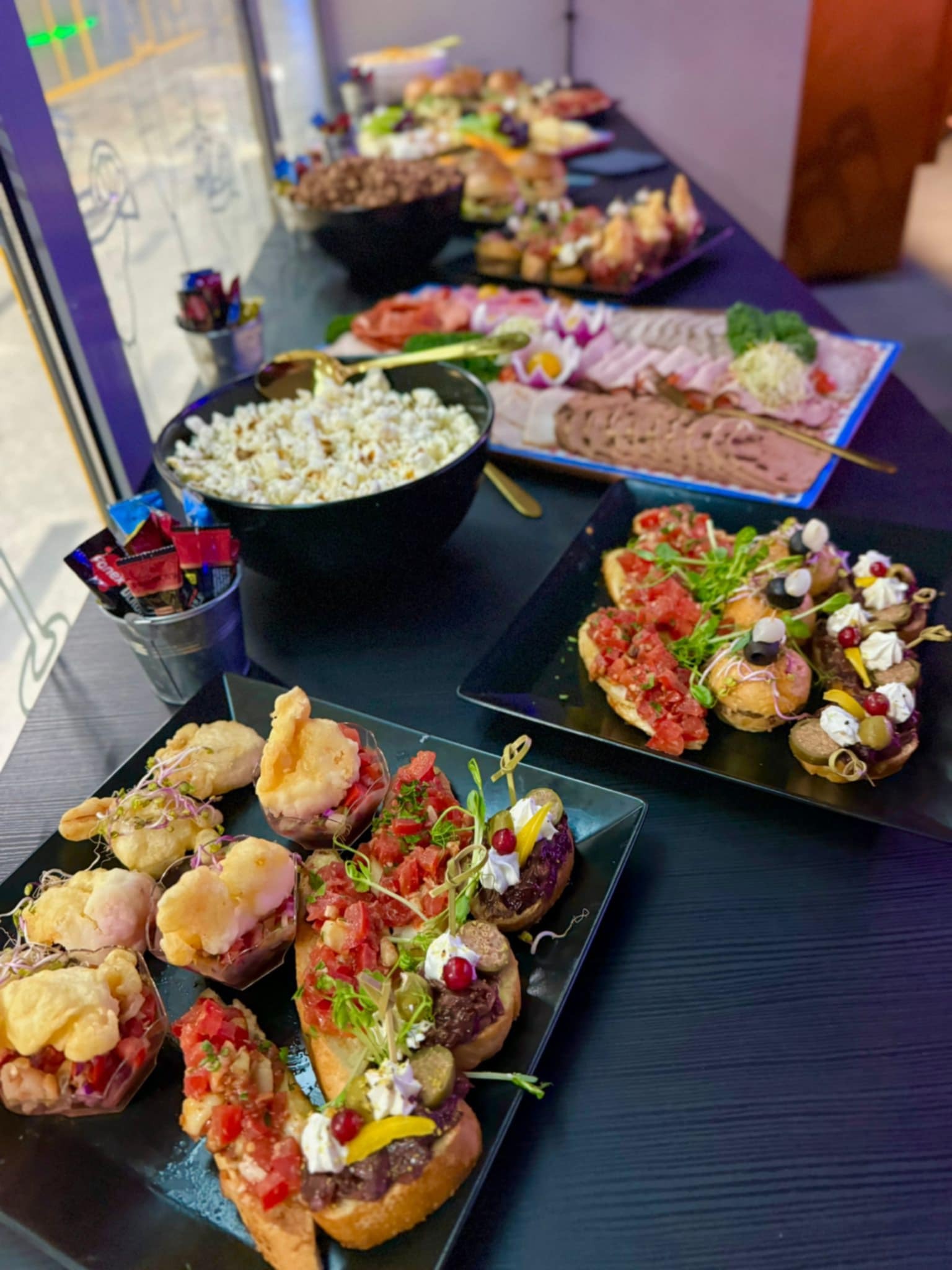 Stół cateringowy z różnorodnymi przekąskami: bruschetta z pomidorami, mini kanapeczki z kremem i oliwkami, smażone krewetki w cieście oraz miska popcornu, uzupełniona o wędliny i sery na półmiskach...