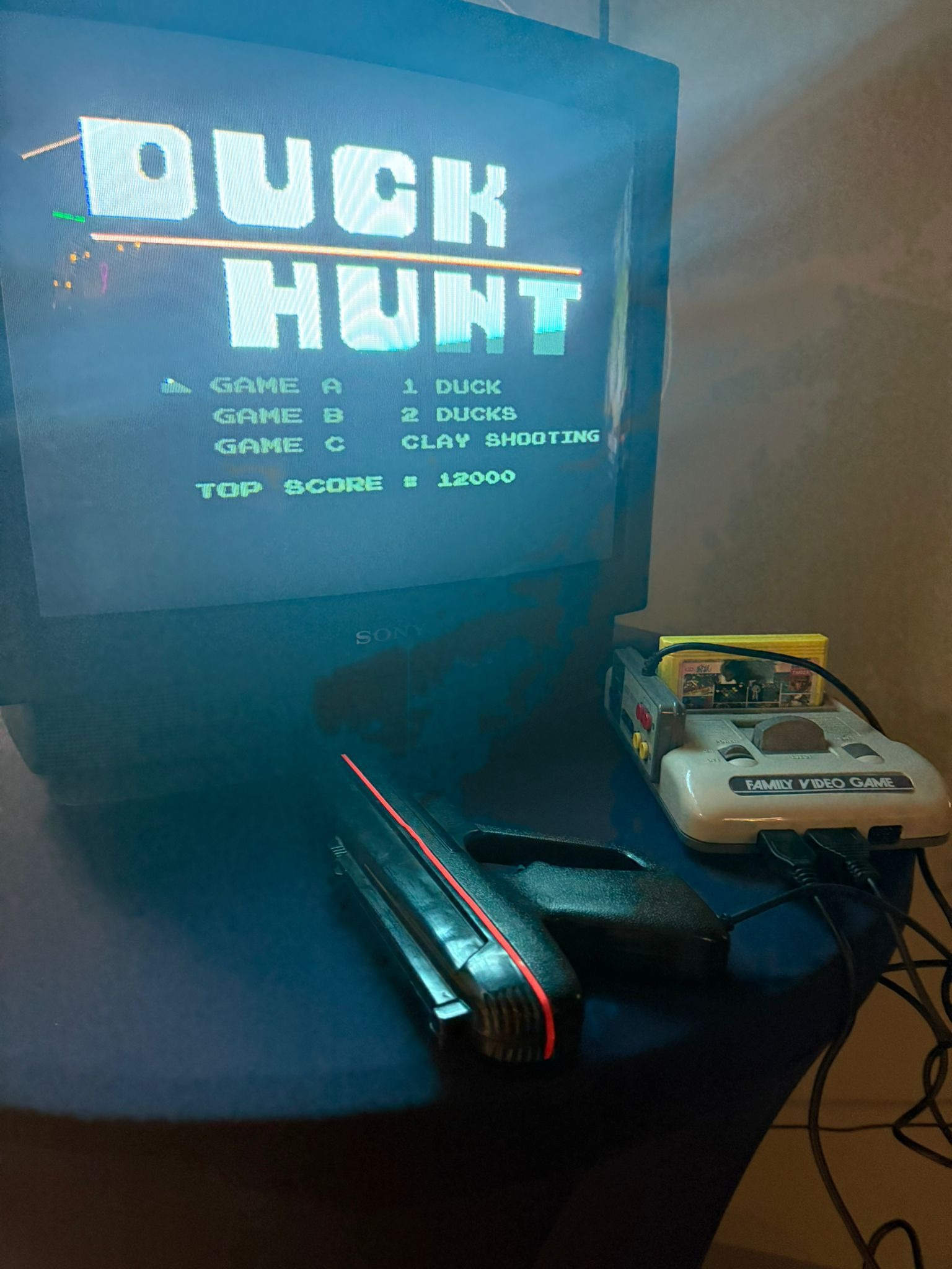 Retro gaming setup featuring a Sony CRT television displaying the title screen of Duck Hunt, accompanied by the console and light gun controller on a blue surface.