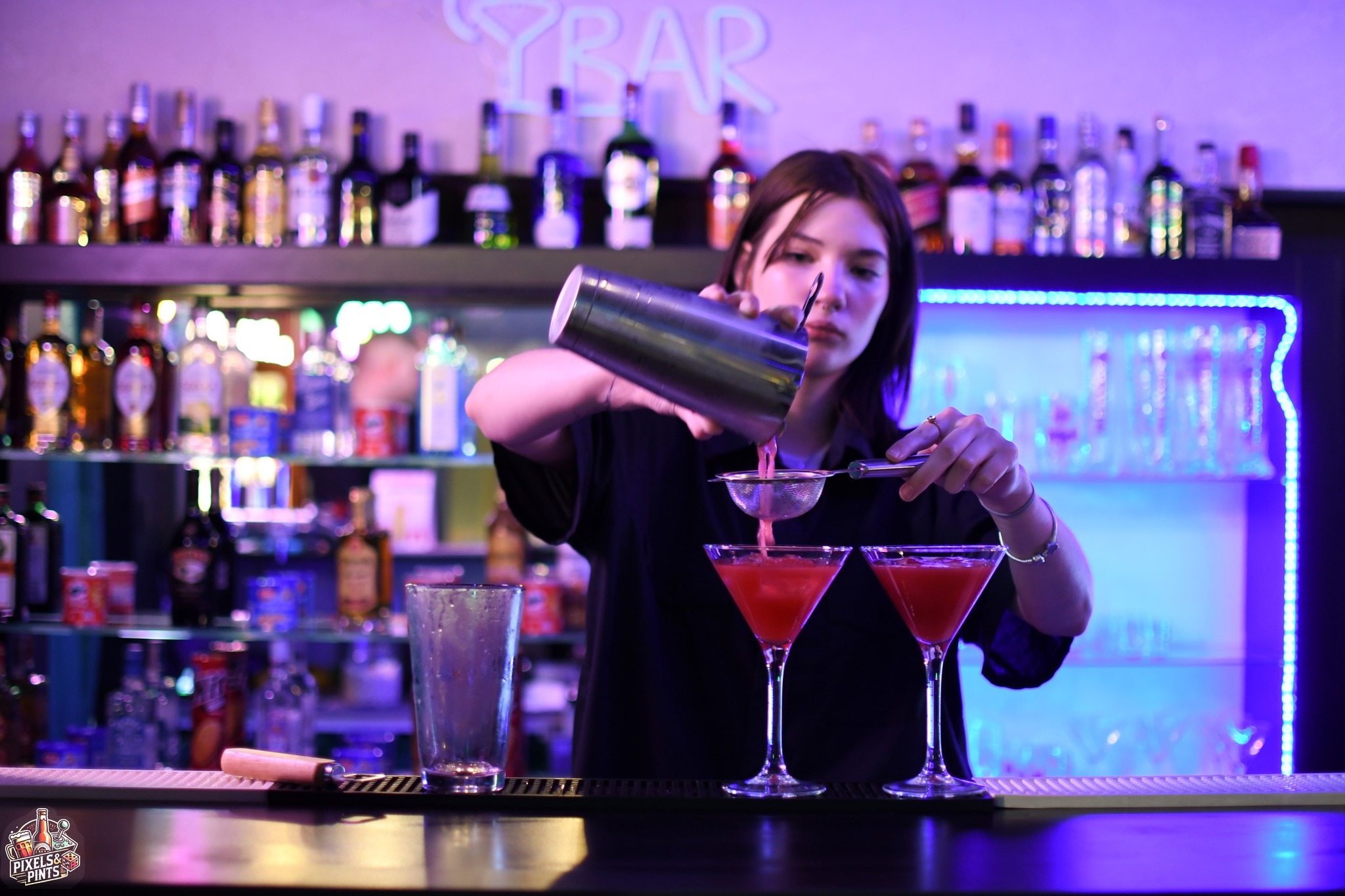 Barmanka przelewa różowy koktajl z shakera przez sitko do kieliszka martini na barze z neonowym napisem i podświetlonymi półkami z alkoholem w tle.