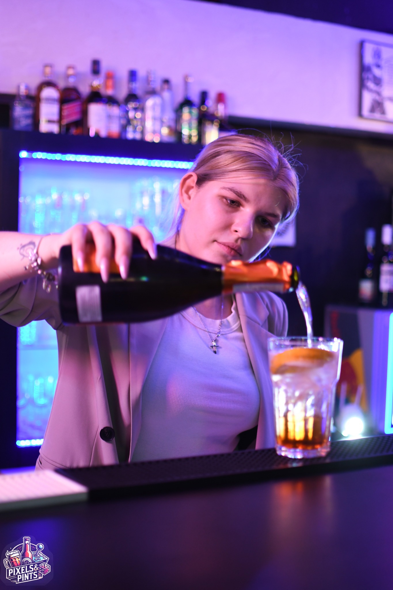 Barmanka w beżowej marynarce nalewa musujący napój z butelki do szklanki z lodem i pomarańczą przy barze podświetlonym niebieskim światłem.