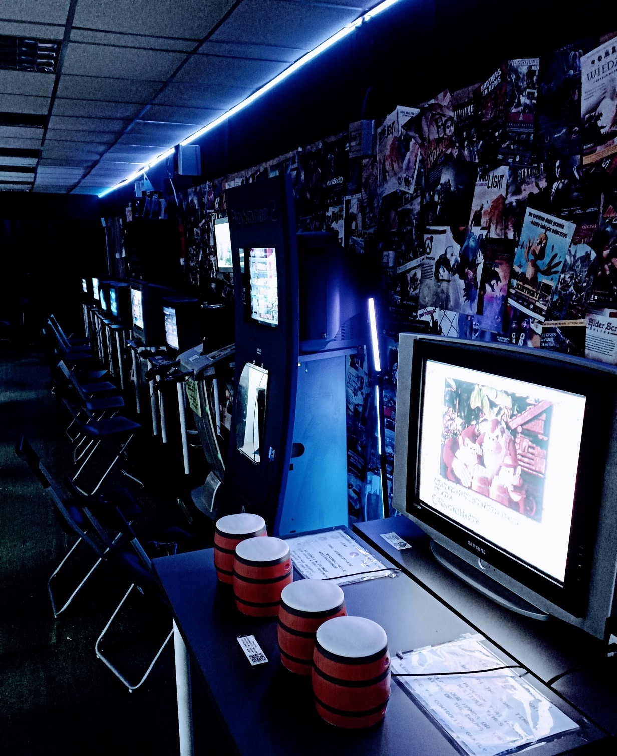 Sala gier z rzędem monitorów, plakatami na ścianach i dekoracjami w postaci małych bębenków. Oświetlenie neonowe podkreśla klimat retro.