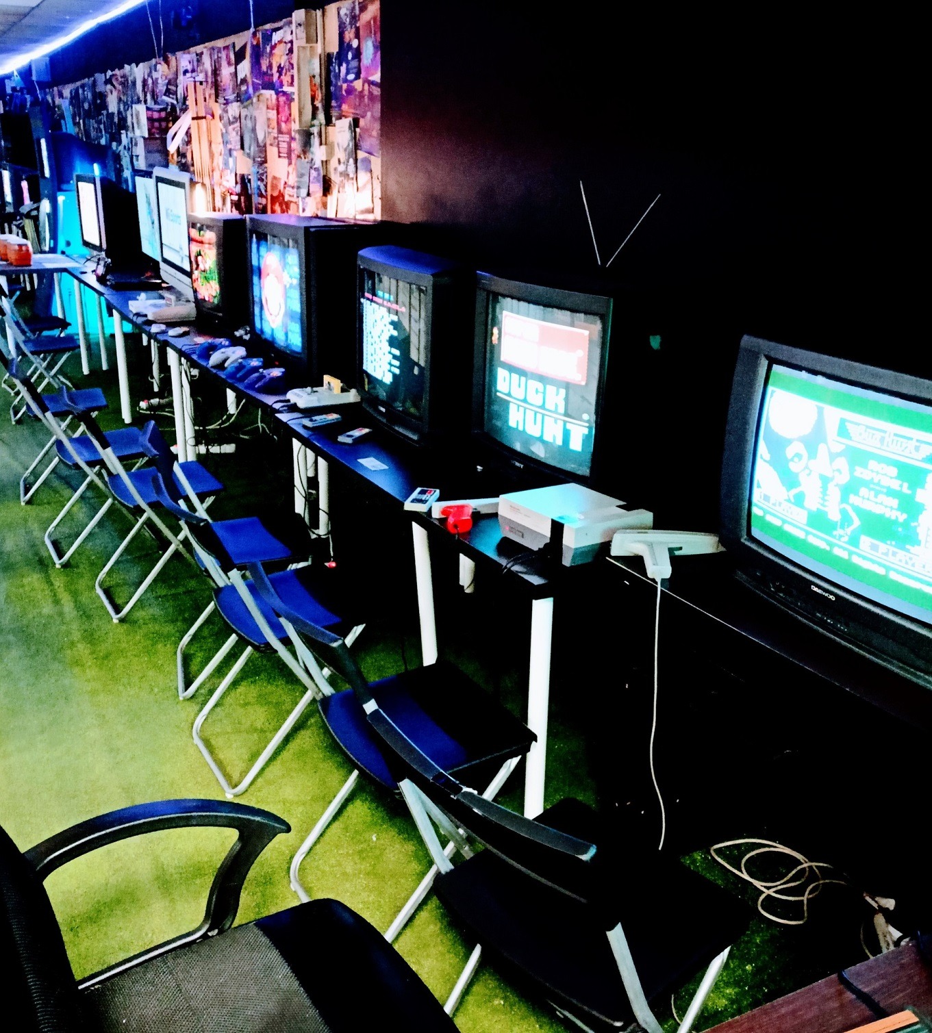 Sala gier retro z rzędem telewizorów CRT, konsolami NES i krzesłami. Ściana ozdobiona plakatami gier, tworzy klimat imprezy tematycznej z grami wideo.