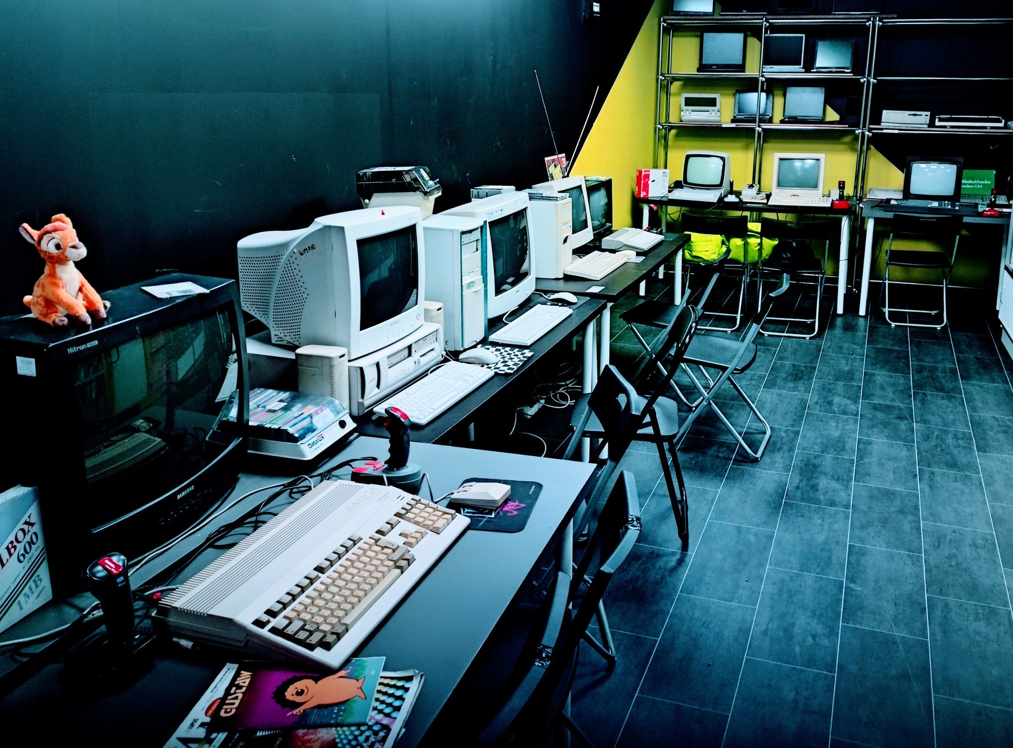 Sala z rzędem komputerów retro i monitorów CRT na biurkach, pluszowy Bambi na monitorze, czarno-żółta ściana, podłoga w płytkach. Klimat retro gamingu.