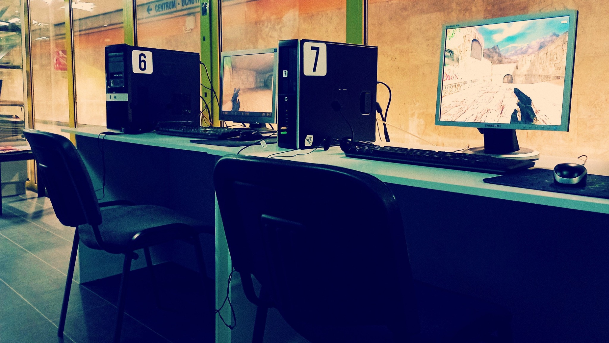Sala z komputerami oznaczonymi numerami 6 i 7, na ekranach widoczna gra Counter-Strike. Krzesła stoją przed stanowiskami. Widoczne okno z napisem.