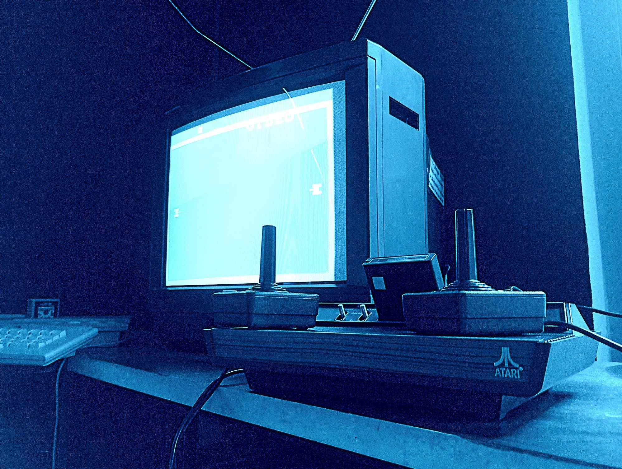Atari z grą na ekranie monitora, dwa joysticki na konsoli. Całość w odcieniach niebieskiego, tworzy klimat retro. Widoczna klawiatura z boku.