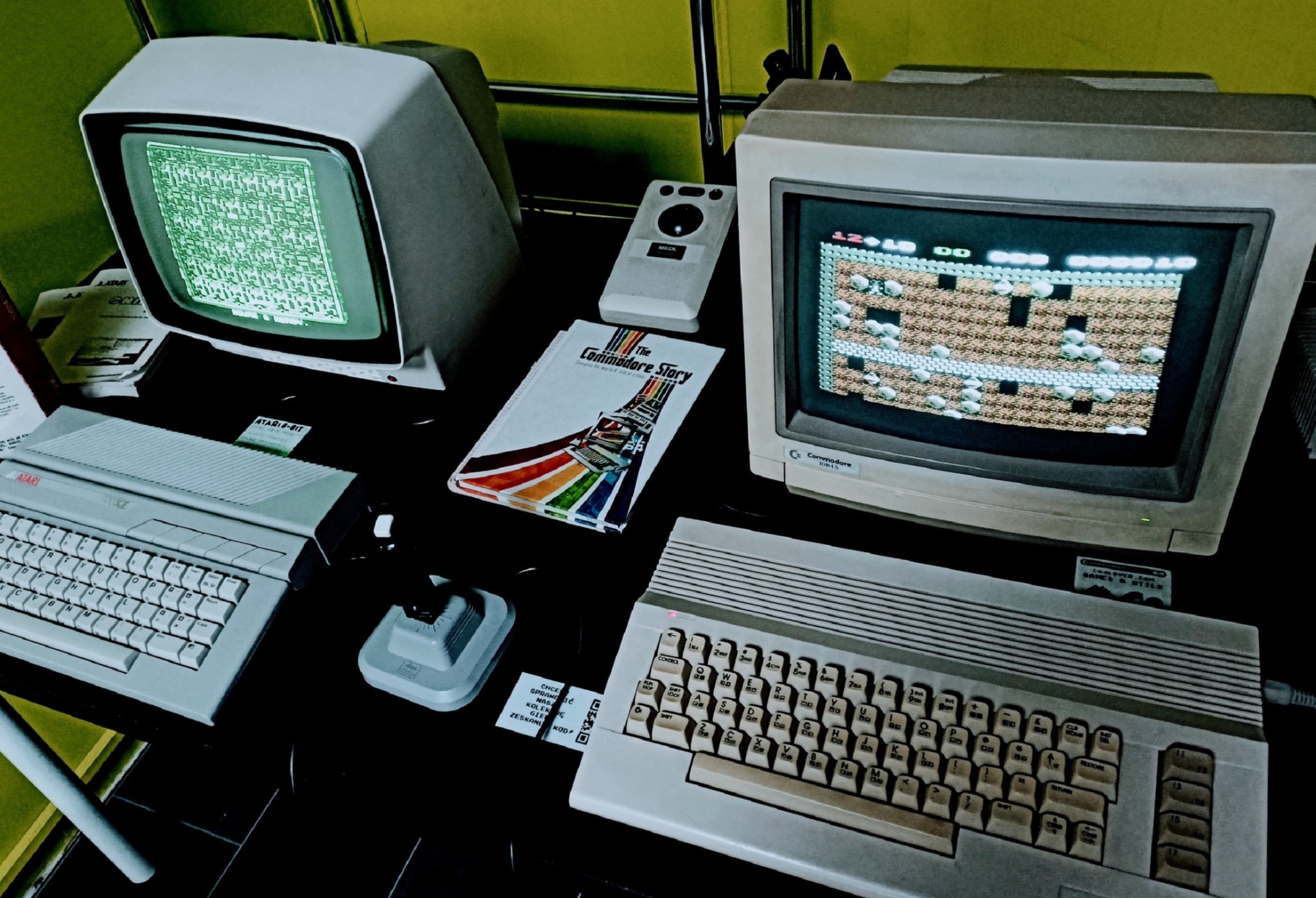 Retrogaming: Dwa komputery Atari i Commodore z monitorami, joystickiem i książką o Commodore. Ekrany wyświetlają gry z lat 80. w stylu retro.