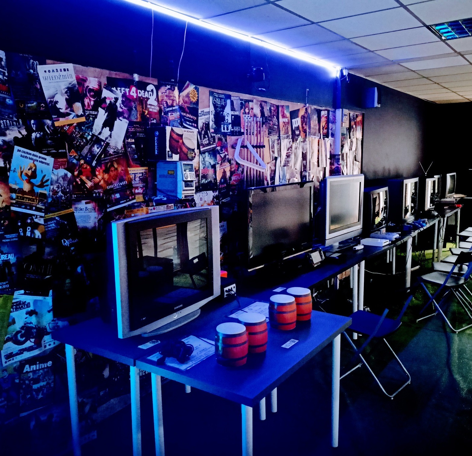 Sala gier z rzędami monitorów CRT, plakatami gier na ścianie i bębnami do gry na stolikach, oświetlona niebieskim światłem.