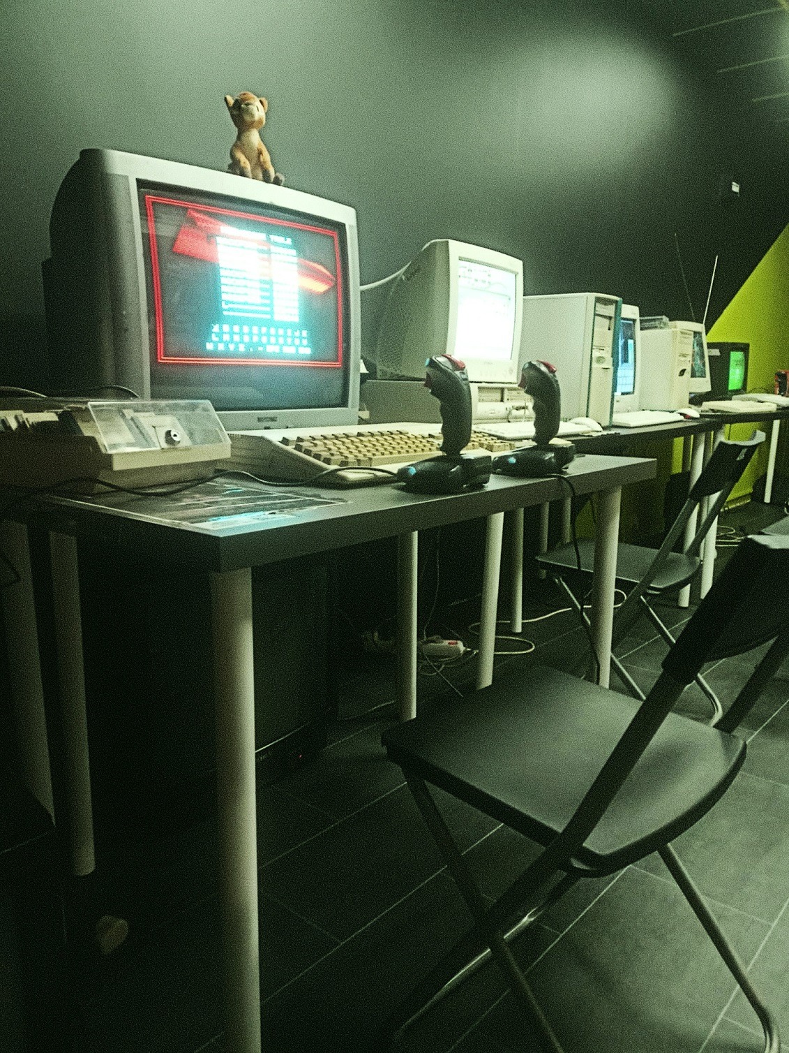 Rząd retro komputerów z joystickami na stołach, ustawionych w ciemnym pomieszczeniu z zielonym akcentem. Na monitorze pierwszego komputera widoczna grafika w stylu retro.