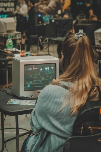 Kobieta gra w grę retro na starym monitorze podczas wydarzenia z grami. W tle widoczni inni gracze i monitory. Atmosfera nostalgii i rywalizacji.