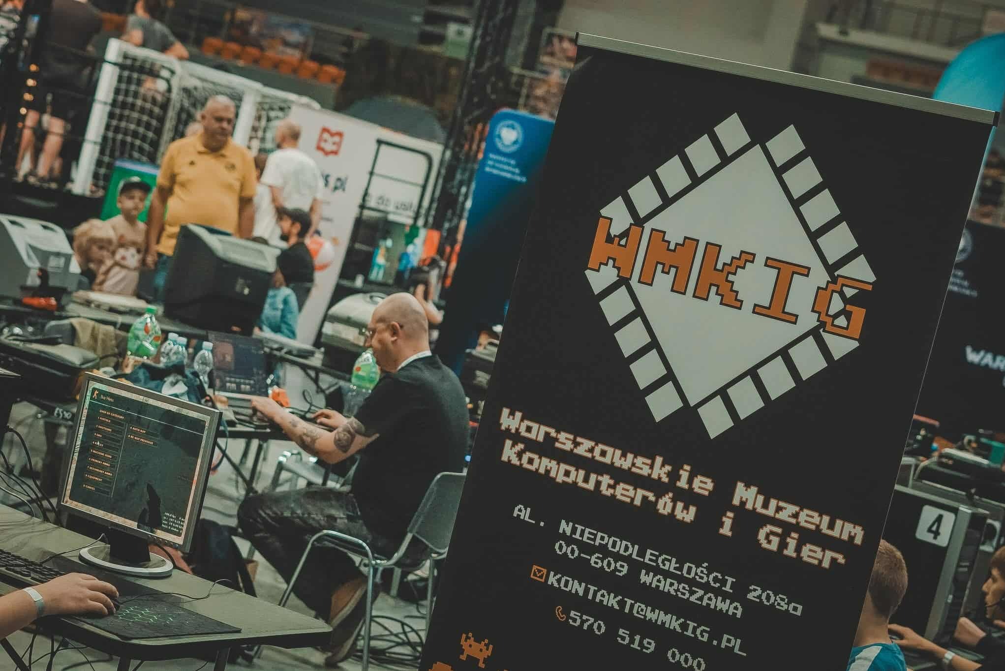 Stoisko Warszawskiego Muzeum Komputerów i Gier na evencie, z osobami grającymi w gry na komputerach. Widoczny baner z logo muzeum w stylu pixel art.