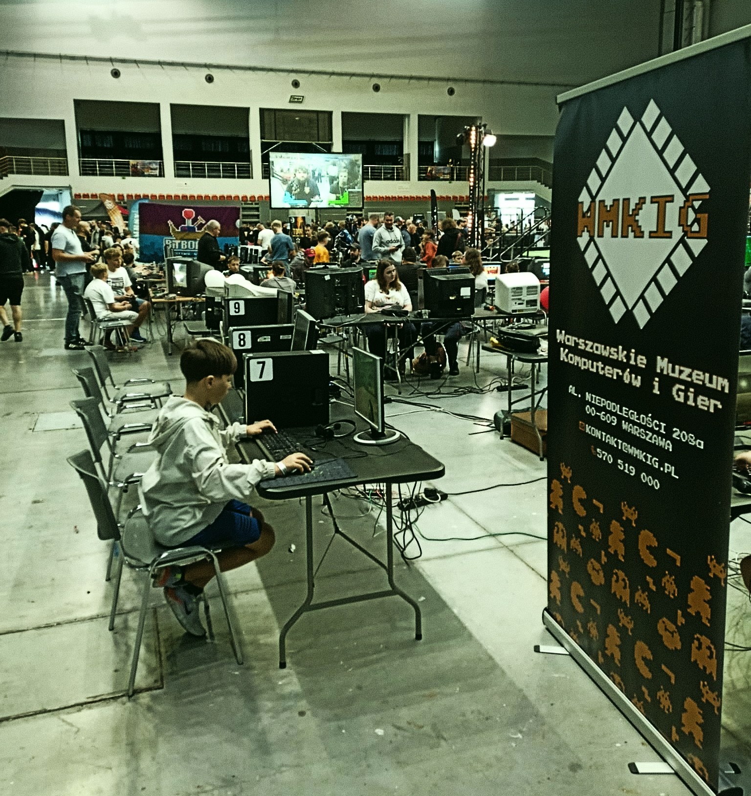 Chłopiec gra na komputerze na evencie Warszawskiego Muzeum Komputerów i Gier, banner z logo, tłum ludzi w tle, numery stanowisk.
