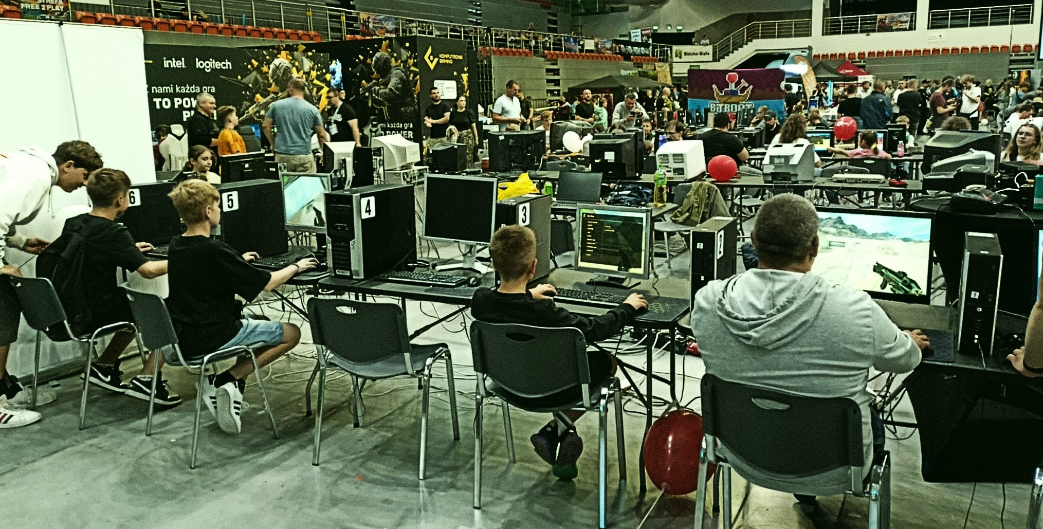 Grupa osób uczestniczy w turnieju gier komputerowych, siedząc przy stanowiskach z monitorami. W tle widoczna publiczność i banery reklamowe sponsorów wydarzenia.