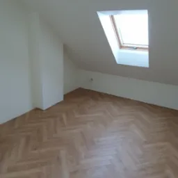 Jasne, wykończone poddasze z oknem dachowym i parkietem ułożonym w jodełkę. Świeżo pomalowane ściany i sufit. Minimalistyczny wystrój, brak mebli.