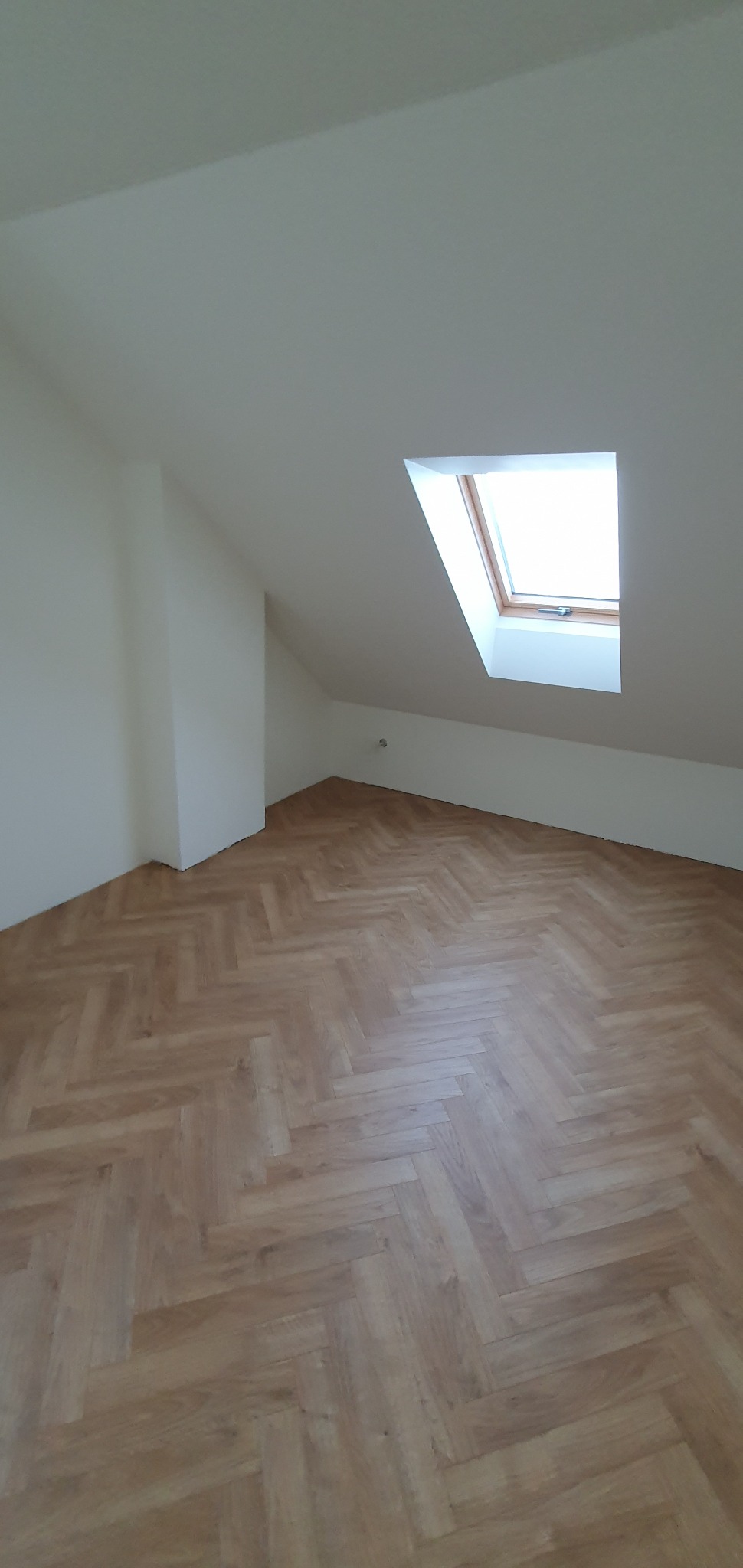 Jasne, wykończone poddasze z oknem dachowym i parkietem ułożonym w jodełkę. Świeżo pomalowane ściany i sufit. Minimalistyczny wystrój, brak mebli.