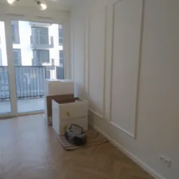 Wnętrze mieszkania w trakcie prac wykończeniowych: białe ściany z dekoracyjnymi listwami, podłoga w jodełkę, niezmontowane białe szafki w kartonach i radio na podłodze, widok z okna na budynek...