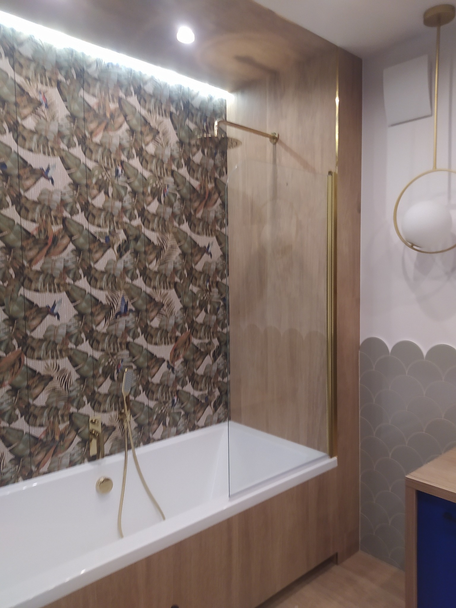 Nowoczesna łazienka z wanną i parawanem nawannowym, wykończona panelami imitującymi drewno, wzorzystą tapetą i złotą armaturą, oświetlona lampą wiszącą.
